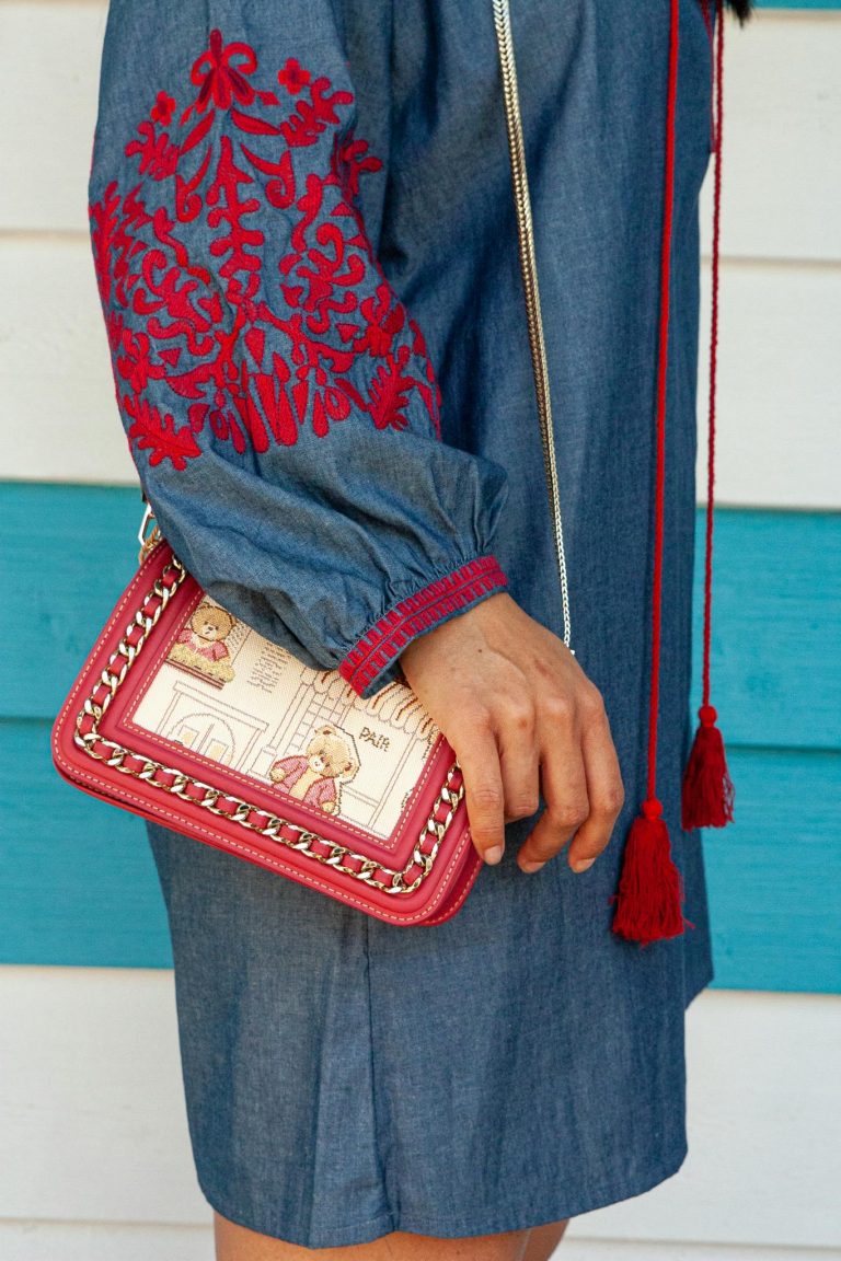A person wearing a denim dress with red embroidery, holding a trendy handbag.