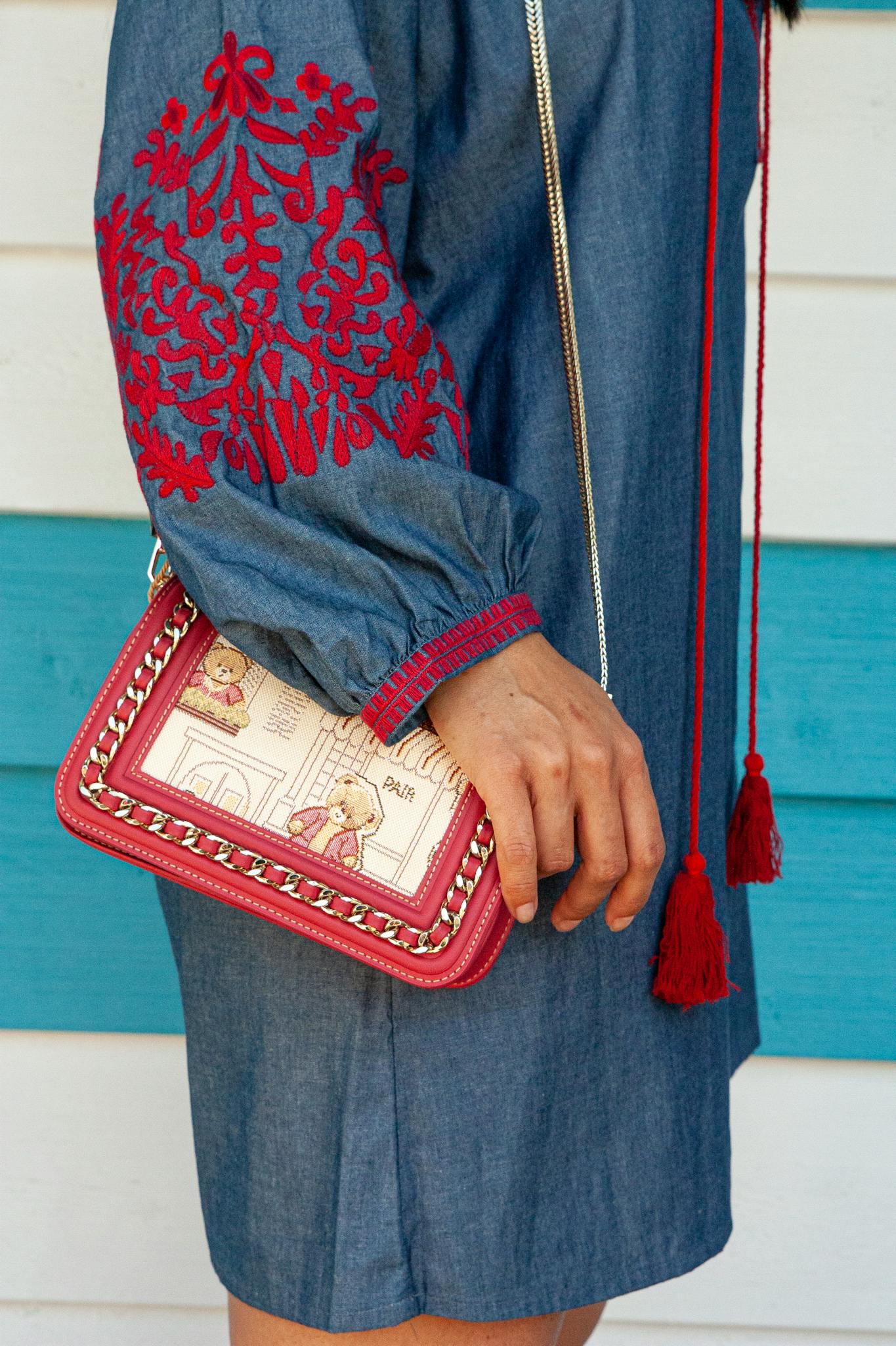 A person wearing a denim dress with red embroidery, holding a trendy handbag.